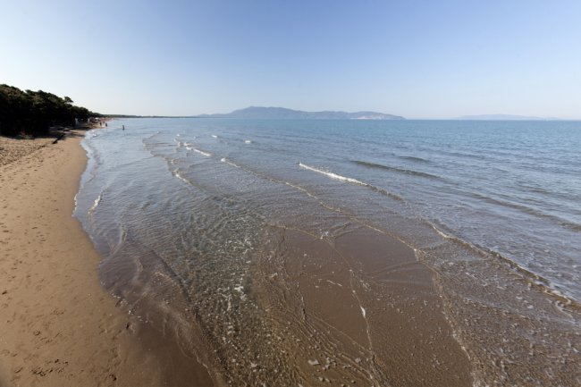Riva del mare con onde leggere, cielo sereno e colline in lontananza.