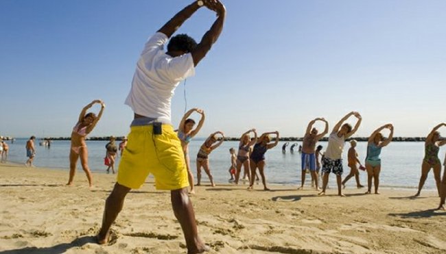 Persone fanno esercizi di stretching su una spiaggia davanti al mare.