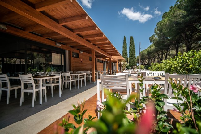 Portico di legno con tavoli e sedie bianche, circondato da alberi e cespugli fioriti.