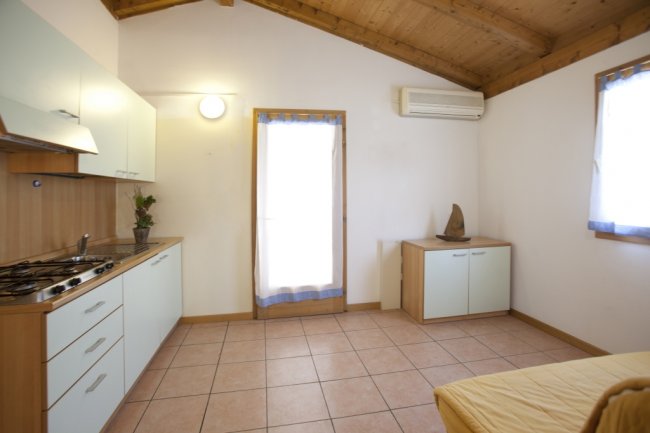 Cucina con mobili chiari, piano cottura e piccola credenza, illuminata da una finestra.