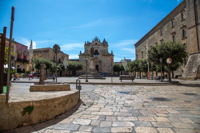 Piazza con chiesa storica, panchine e pavimentazione in pietra sotto un cielo blu.