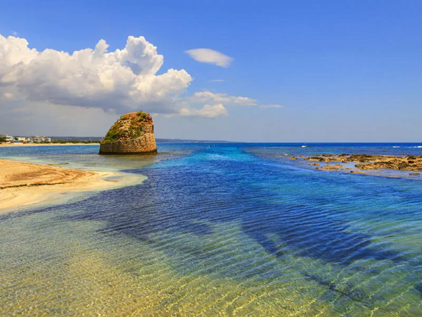 Spiaggia con mare limpido e una formazione rocciosa. Cielo azzurro con nuvole.