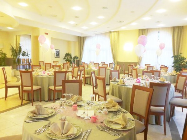 Sala decorata per festa con tavoli rotondi, tovaglie eleganti e palloncini rosa e bianchi.