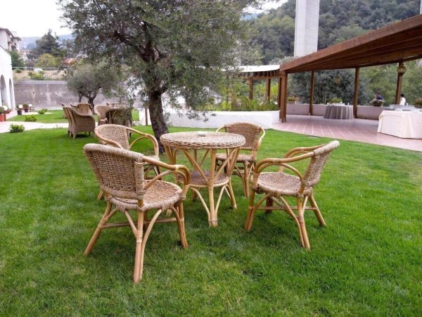 Tavolo con sedie in vimini su un prato verde, vicino a un ulivo e una struttura in legno.