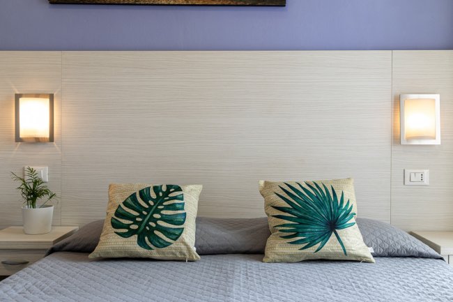 Letto con cuscini a tema foglie tropicali, lampade moderne e un piccolo vaso di piante.