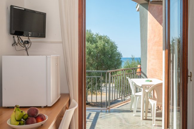 Tavolo e sedie sul balcone con vista mare, TV e frigorifero accanto a una ciotola di frutta.
