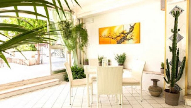 Area interna luminosa con tavolo, sedie bianche e piante. Quadro giallo sulla parete.