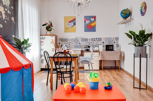 Spazio colorato con giochi per bambini, tavolo, sedie e angolo cucina. Decorazioni vivaci e piante.