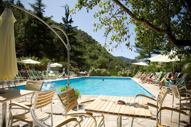 Una piscina all'aperto circondata da sedie e alberi, con persone rilassate sotto gli ombrelloni.