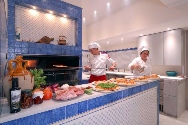 Due chef preparano pizze in una cucina con rivestimenti blu, con ingredienti freschi sul bancone.