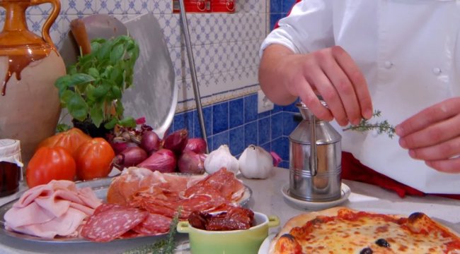 Un cuoco prepara una pizza circondato da salumi, verdure e spezie.