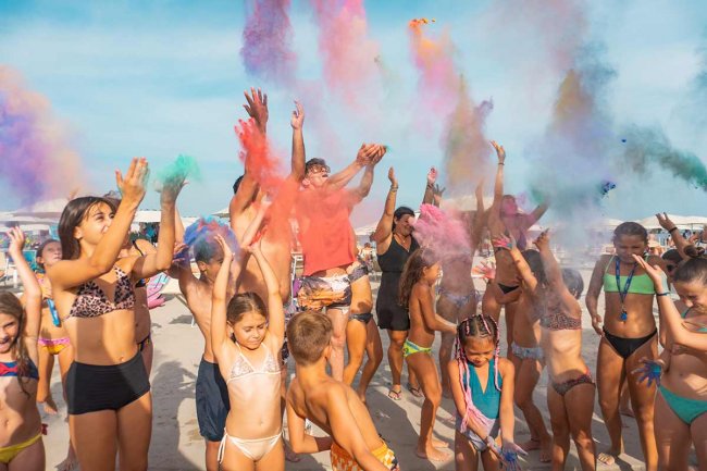 Gruppo di persone su una spiaggia lancia polveri colorate in aria, creando un'atmosfera festosa.