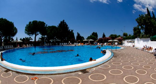 Ampia piscina con bagnanti e sdraio, circondata da alberi.