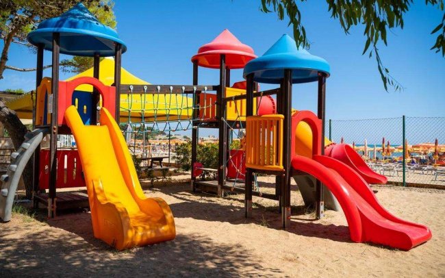 Un parco giochi colorato con scivoli rossi e gialli su sabbia, sotto cieli blu.