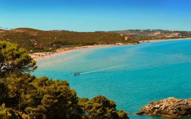 Spiaggia panoramica con acque turchesi e colline verdi in lontananza.