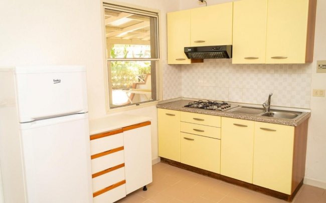 Cucina con mobili gialli, piano cottura, frigo bianco e finestra.